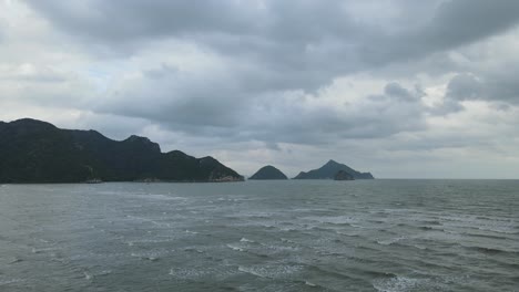 A-steady-aerial-footage-of-waves-rushing-to-the-shore,-mountains,-islands-in-the-horizon,-beautiful-gray-clouds