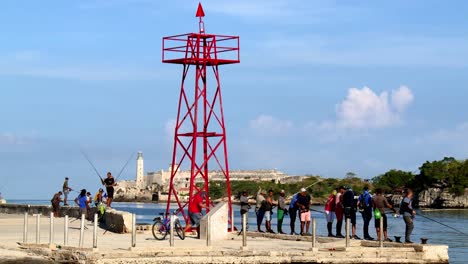 Menschen,-Die-Vom-Dock-Mit-Dem-Leuchtturm-Faro-Castillo-Del-Morro-Im-Hintergrund-In-Havanna,-Kuba,-Fischen