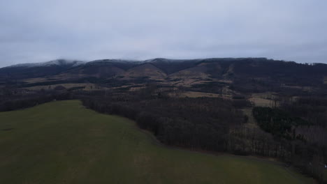 Fliegen-Sie-über-Die-Landschaft-Mit-Dem-Wald-Unter-Ihnen-Im-Hintergrund-Der-Schneebedeckten-Hügel