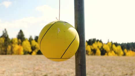 Gelber-Tetherball,-Der-An-Einem-Seil-Mit-Herbstlaub-Auf-Verschwommenem-Hintergrund-An-Einer-Stationären-Metallstange-Aufgehängt-Ist