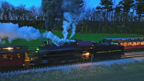 A-Drone-Slightly-Ahead-and-Parallel-Night-View-of-a-Steam-Passenger-Train-Stopped-as-Another-Steam-Train-Passes-Blowing-Lots-of-Smoke-Seeing-the-Lights-in-the-Coaches