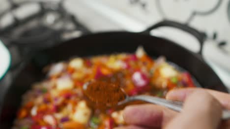 Close-up-focus-on-a-spoon-with-red-spice-put-into-vegetable-mix-inside-hot-frying-pan