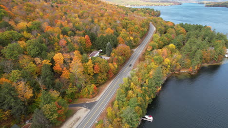 Aerial-View-of-Lonely-Dar-Car-Moving-on-Road-by-Sunapee-Lake-and-Colorful-Forest-in-Vivid-Autumn-Foliage,-New-Hampshire-USA,-Tracking-Drone-Shot