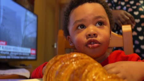 Two-year-old-black-baby-tries-to-eat-a-big-croissant-by-himself-at-home-seated-next-to-the-TV