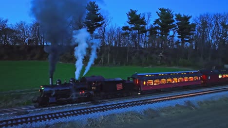 A-Drone-Slightly-Ahead-and-Parallel-Night-View-of-a-Steam-Passenger-Train-Stopped-in-Farmlands-Blowing-Lots-of-Smoke-Seeing-the-Lights-in-the-Coaches