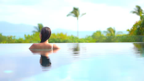 A-woman-in-a-swimming-pool-with-her-back-to-the-camera-looks-out-over-the-tropical-ocean-view