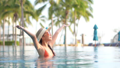 Asian-Woman-in-Orange-Swimsuit-Raising-Arms-up-inside-Swimming-Pool-Water-on-Lush-Tropical-Palms-Background-in-Bali-at-Sunset,-slow-motion
