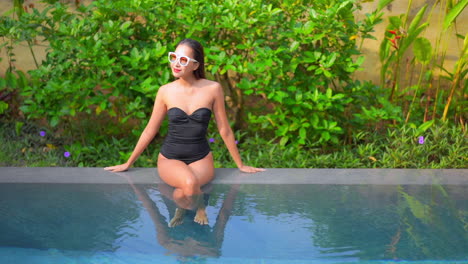 Asian-woman-sitting-on-swimming-pool-edge-with-black-monokini-and-white-sunglasses,-legs-crossed-with-green-bushes-on-background---pulling-focus-shot