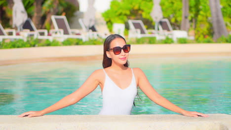 A-young-woman-standing-in-a-resort-swimming-pool-reaches-up-to-adjust-her-sunglasses
