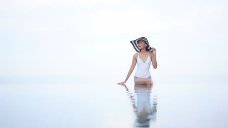 Asian-woman-in-a-white-bathing-suit-and-floppy-striped-sunhat-sits-on-the-edge-of-the-high-rooftop-infinity-pool-with-feet-in-the-water-together-with-body-reflection-in-water---copy-space-dream-look