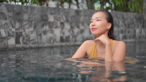 Dreaming-Asian-woman-leaning-chin-on-hand-at-the-edge-of-swimming-pool-and-looking-aside-smiling,-Thailand-resort,-face-closeup-slow-motion