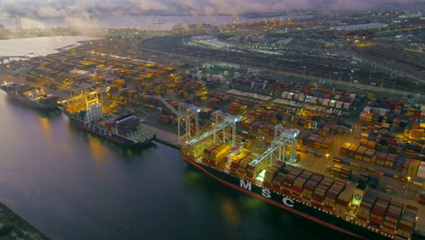 Oakland-port-container-ships-unloading-cargo-illuminated-at-night-aerial-view-rising-back