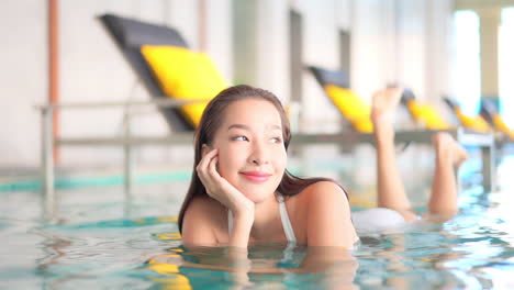 Attractive-hot-Asian-Woman-Lying-Down-on-Her-Belly-in-Shallow-Swimming-Pool-Water-in-Tropical-Hotel-Looking-Aside-Smiling-While-Moving-Legs-Up-and-Down,-slow-motion