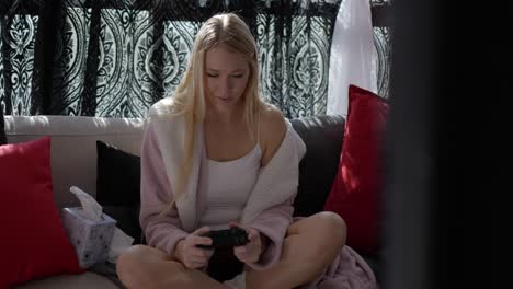 Beautiful-blonde-white-caucasian-girl-women-sitting-on-couch-in-front-of-windows-in-pink-bath-robe-resting-relaxing-at-home-playing-video-games-with-controller-in-hand-smiling-looking-straight-at-tv