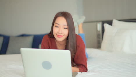 Asian-woman-checking-bitcoin-price-on-laptop,-suddenly-showing-a-winning-expression-and-Yes-gesture-while-lying-down-on-bed