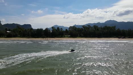 A-Jet-Ski-silhouette-speeding-to-the-right-diagonally-to-the-shore,-beachfront,-mountains-at-the-background,-hot-afternoon,-Dolphin-Bay,-Sam-Roi-Yot-National-Park,-Prachuap-Khiri-Khan,-Thailand