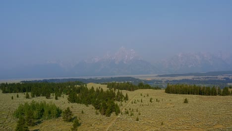 Aerial-drone-landscape-nature-dolly-in-shot-of-the-majestic-Grand-Tetons-National-Park-mountain-range-with-a-valley-of-brush-and-pine-trees-below-on-a-warm-hazy-summer-day-in-Wyoming,-USA