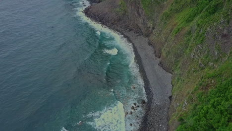 Aerial-panning-up-to-oceanside-waterfall-from-mountains,-Madeira-Portugal