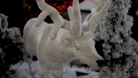 White-plush-reindeer-Christmas-ornament-with-snowed-artificial-trees-behind,-Close-up-shot