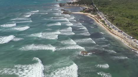 Aerial-drone-shot-of-giant-waves-reaching-sandy-beach-of-Riviera-Maya-in-Tulum---Cars-driving-on-coastline-road---Coral-reef-in-transparent-Caribbean-Sea-Water