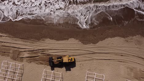 Aerial-top-down-shot-of-Digger-grabbing-and-transporting-sand-on-beach-beside-waves-of-ocean---4K