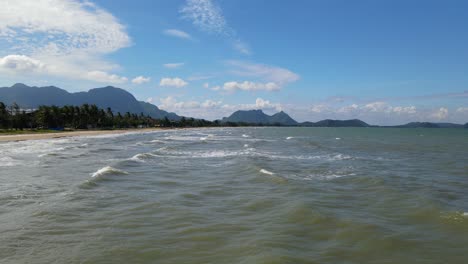 Steady-aerial-footage-of-the-waves-moving-towards-the-shore,-the-beach,-lovely-mountains-and-sky