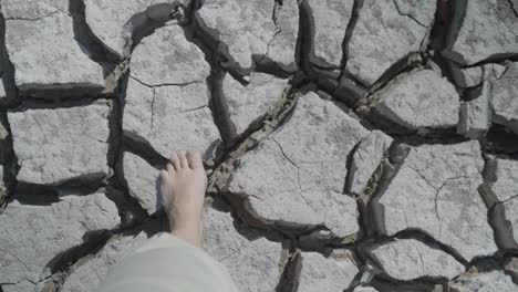 Point-of-view-looking-down-at-bare-feet-walking-across-dry,-cracked-land