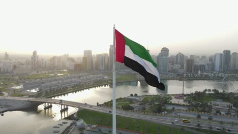 La-Bandera-De-Los-Emiratos-árabes-Unidos-Ondeando-En-El-Aire,-El-Cielo-Azul-Y-El-Desarrollo-De-La-Ciudad-En-El-Fondo,-El-Símbolo-Nacional-De-Los-Emiratos-árabes-Unidos-Sobre-La-Isla-De-La-Bandera-De-Sharjah