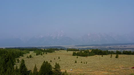 Aerial-drone-landscape-nature-tilting-up-shot-of-the-Grand-Tetons-National-Park-mountain-range-with-a-valley-of-brush-and-pine-trees-and-small-campground-below-on-a-warm-hazy-summer-day-in-Wyoming