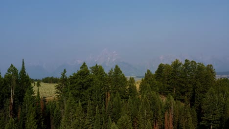 Aerial-drone-landscape-nature-shot-passing-over-the-tops-of-pine-trees-and-revealing-the-Grand-Tetons-National-Park-mountain-range-with-a-valley-of-brush-and-pine-trees-behind-in-Wyoming,-USA
