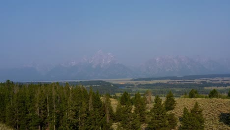 Aerial-drone-landscape-nature-shot-passing-over-the-tops-of-pine-trees-with-the-Grand-Tetons-National-Park-mountain-range-with-a-valley-of-brush-and-pine-trees-behind-in-Wyoming,-USA