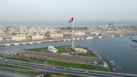 Die-Flagge-Der-Vereinigten-Arabischen-Emirate-Weht-In-Der-Luft,-Der-Blaue-Himmel-Und-Die-Stadtentwicklung-Im-Hintergrund,-Das-Nationale-Symbol-Der-Vae-über-Der-Flaggeninsel-Von-Sharjah