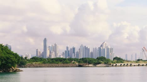 Handheld-shot-of-landscape-with-skycraper-skyline-in-Panama-City's-bay-waterline-viewed-from-Amador's-Causeway-during-a-sunny-summer-day-behind-dense-vegetation-mangrove-and-a-cloudy-blue-sky
