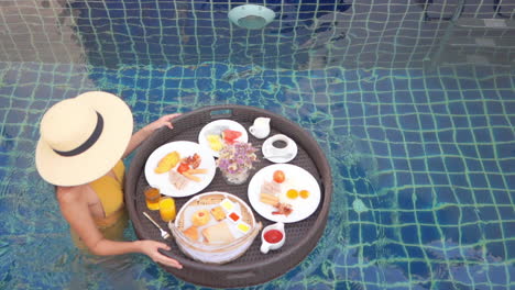 Woman-in-Swimsuit-Walking-in-Pool-Water-With-Floating-Plate-and-Served-Breakfast-in-Luxury-Hotel-Resort,-Slow-Motion