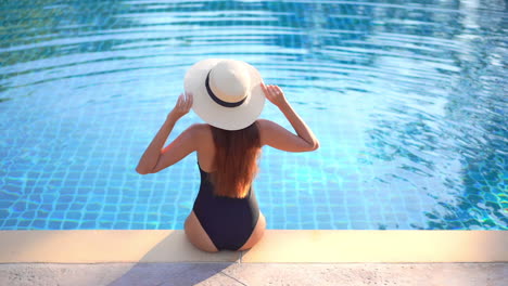 Back-of-Woman-WIth-Perfect-Body-Figure-Sitting-on-Pool-Edge-in-Swimsuit-and-Floppy-Summer-Hat-on-Hot-Sunny-Day,-Full-Frame