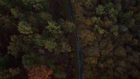 Drone-tracking-of-a-car-with-lights-on-on-a-road-through-the-forest-at-the-end-of-the-day-in-autumn,-Dordogne---France