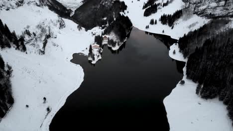 Aerial-flyover-over-lake-Seealpsee-in-Appenzell,-Switzerland-with-a-pan-up-motion-revealing-the-winter-landscape-full-of-snow-and-cliffs