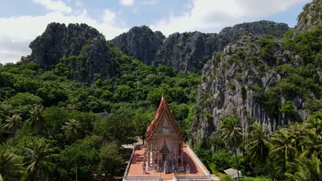 Aerial-footage-towards-the-Buddhist-temple-from-a-high-altitude-revealing-coconuts,-limestone-mountains,-forest-and-sky