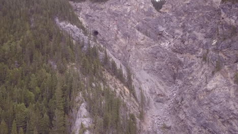 Rocky-mountain-aerial-flies-up-steep-dry-drainage-to-large-cave-mouth