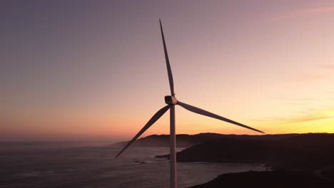 Aerial-drone-close-up-turbine-wind-windmill-during-sunset-with-ocean-cliff-on-the-background,-alternative-renewable-energy