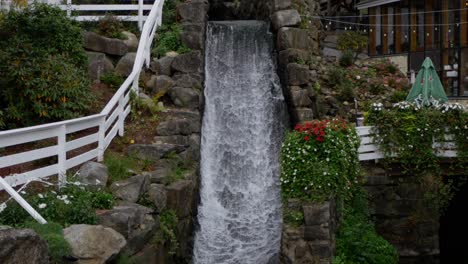 A-small-waterfall-runs-near-a-patio-and-flowers-as-the-camera-tilts-to-its-base