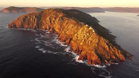 Aerial-front-view-of-the-European-end-of-the-earth-cape-finisterre-Cabo-fisterra-during-sunset,-lighthouse-chapel-on-top-of-rocky-cliff-mountain-formation-in-the-Atlantic-sea-ocean