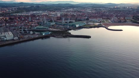 Aerial-view-of-gijon-city-coastline-in-north-Spain-during-sunset,-sand-beach-and-commercial-harbor