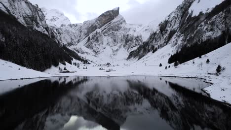 Sobrevuelo-Aéreo-A-Través-Del-Lago-Seealpsee-En-Appenzell,-Suiza,-Rodeado-De-Nieve-En-Un-Día-De-Invierno-Con-Un-Reflejo-De-Los-Picos-Alpstein-En-Las-Aguas-Tranquilas