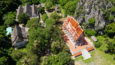 Aerial-footage-sliding-to-the-left-from-a-high-altitude-revealing-this-temple-complex,-limestone-mountains,-coconuts,-Wat-Khao-Daeng,-Khao-Sam-Roi-Yot-National-Park,-Phrachuap-Khiri-Khan,-Thailand
