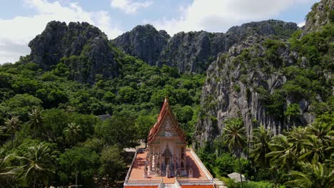 Aerial-footage-from-a-higher-vantage-altitude-towards-this-lovely-temple-revealing-mountains,-coconut-trees-and-forest,-Wat-Khao-Daeng,-Khao-Sam-Roi-Yot-National-Park,-Phrachuap-Khiri-Khan,-Thailand