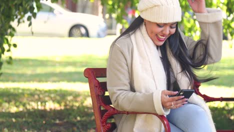 Attractive-girl-sitting-on-a-city-park-bench-texting-during-the-chilly-fall-and-autumn-season