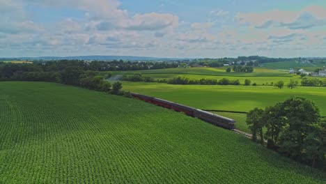 An-Aerial-Traveling-View-of-Corn-Fields-and-Harvesting-Crops,-with-Patches-of-Color-With-a-Passenger-Train-Traveling-Thru-It-on-a-Beautiful-Summer-Day