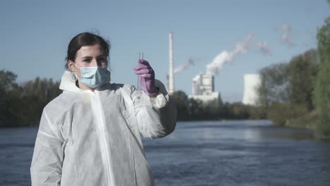 Woman-in-face-mask-and-protective-suit-holding-test-tube-with-water-sample,-smoking-factory-in-background
