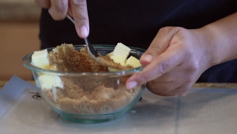 Mixing-brown-sugar-and-butter-with-a-fork-in-a-glass-bowl---black-hands-preparing-treat
